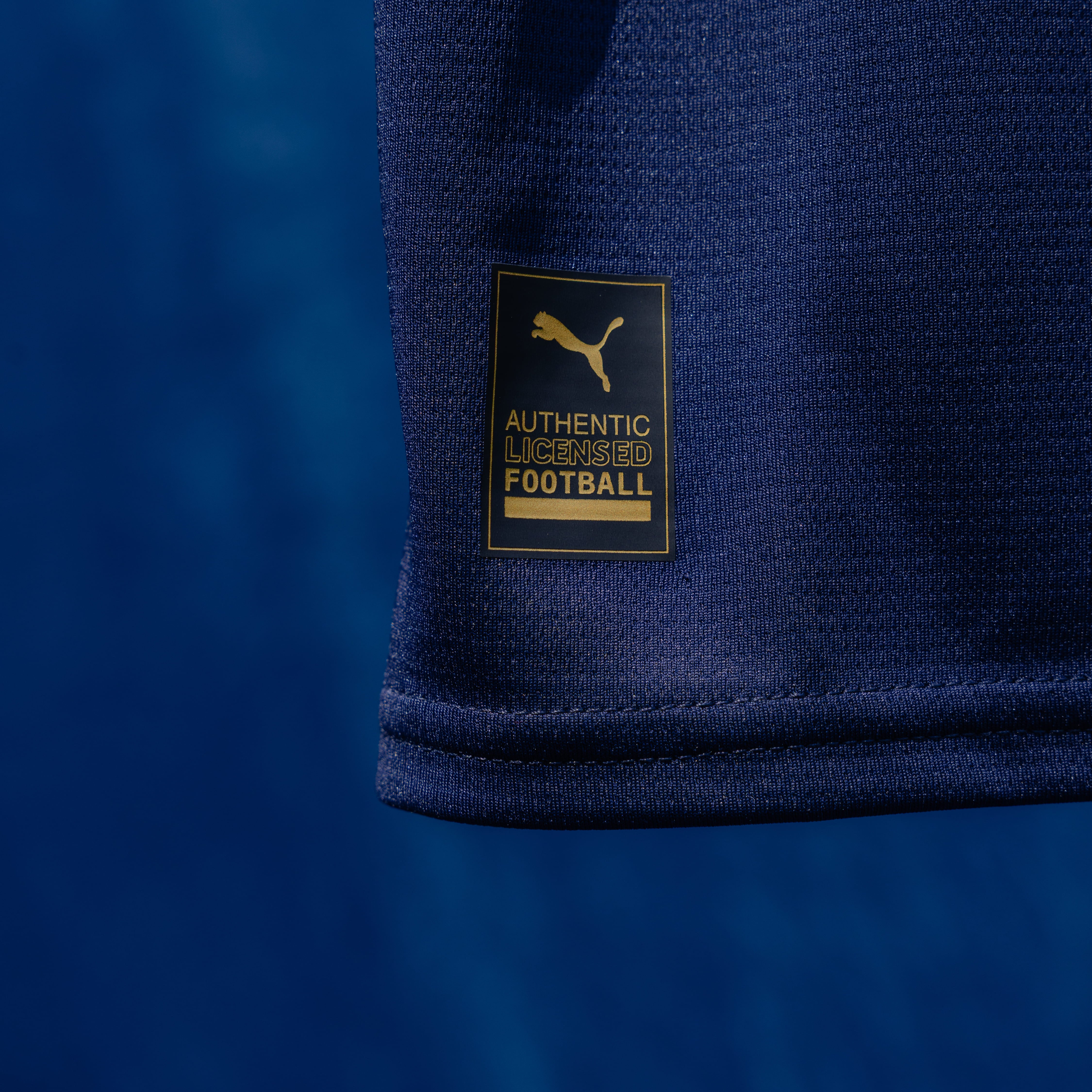 Dettaglio targhetta "AUTHENTIC LICENSED FOOTBALL" con Puma Cat in colore oro su maglia gara Fourth blu navy