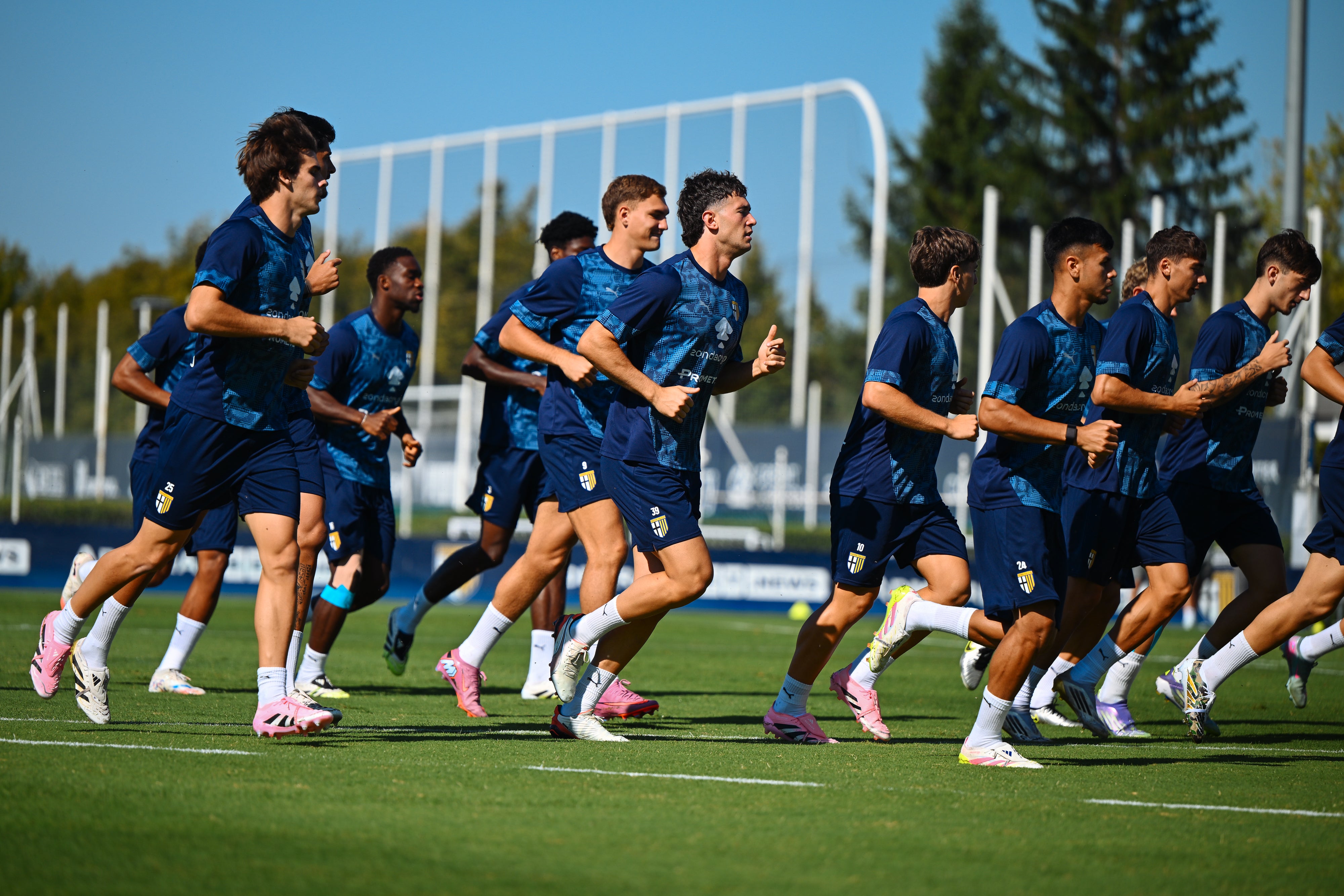 Foto allenamento prima squadra maschile con calciatori mentre corrono indossando maglia mezze maniche e pantaloncino blu.