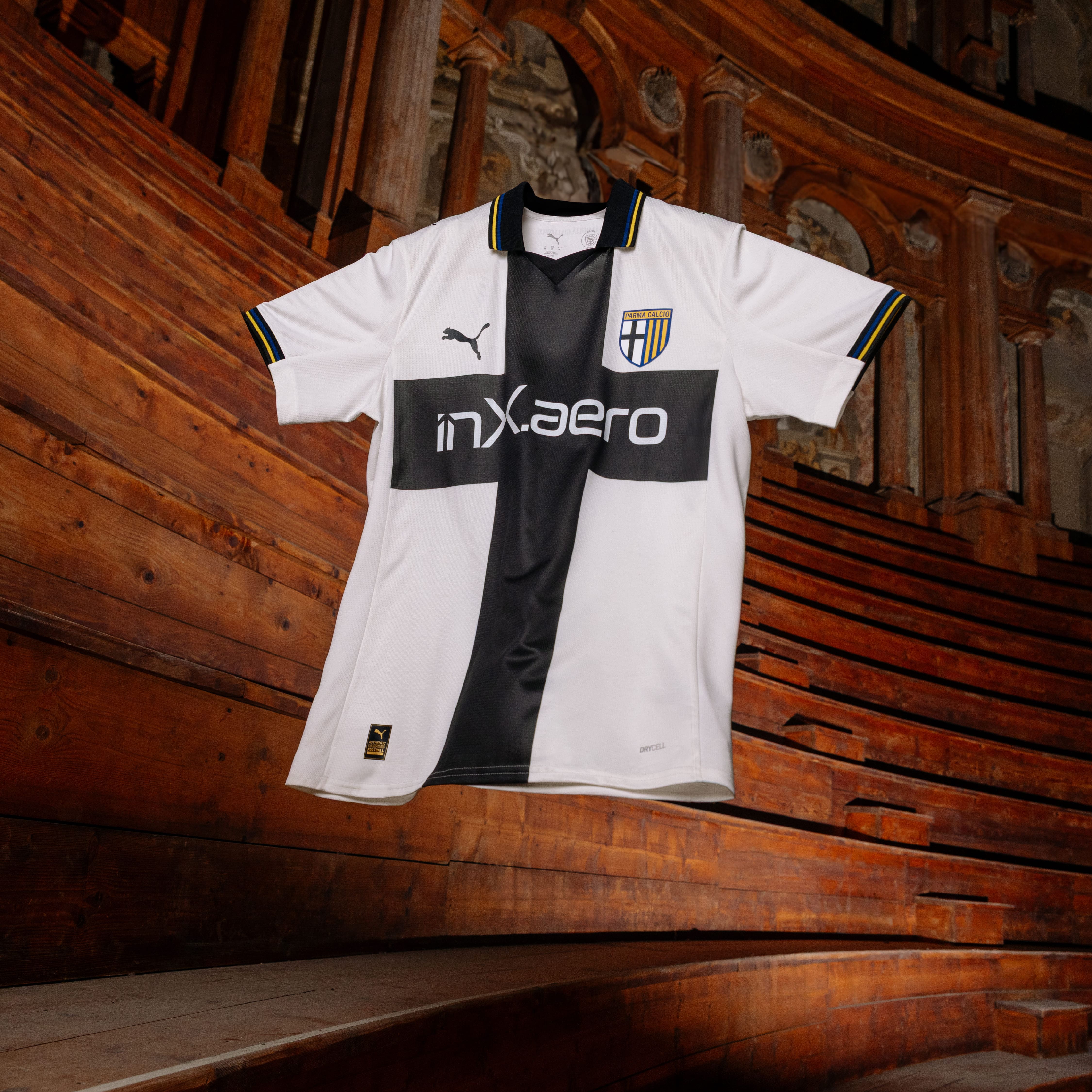 Maglia gara home parma calcio femminile bianca con croce nera e dettagli a righe gialloblu su colletto e maniche sospesa in aria con fondo teatro Farnese