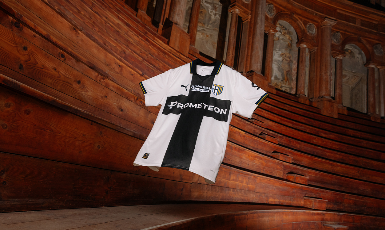 Maglia gara Home Parma Calcio maschile bianca con croce nera e dettagli a righe gialloblu su colletto e maniche sospesa in aria con fondo teatro Farnese