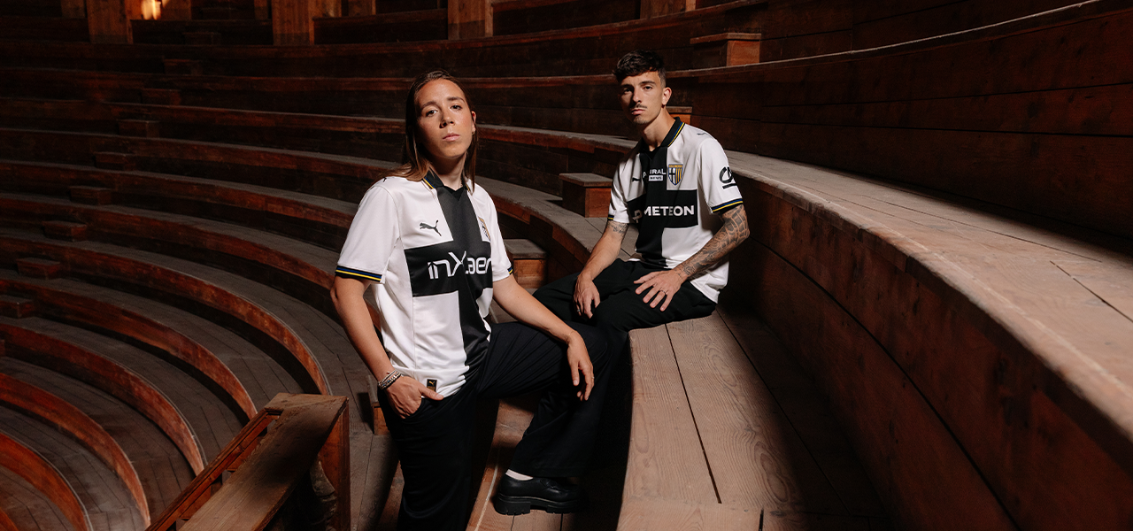 Foto con Ambrosi e Delprato seduti mentre indossano la nuova maglia Home Parma Calcio 2025/26 bianca con croce nera e dettaglio righe gialloblu su colletto e maniche