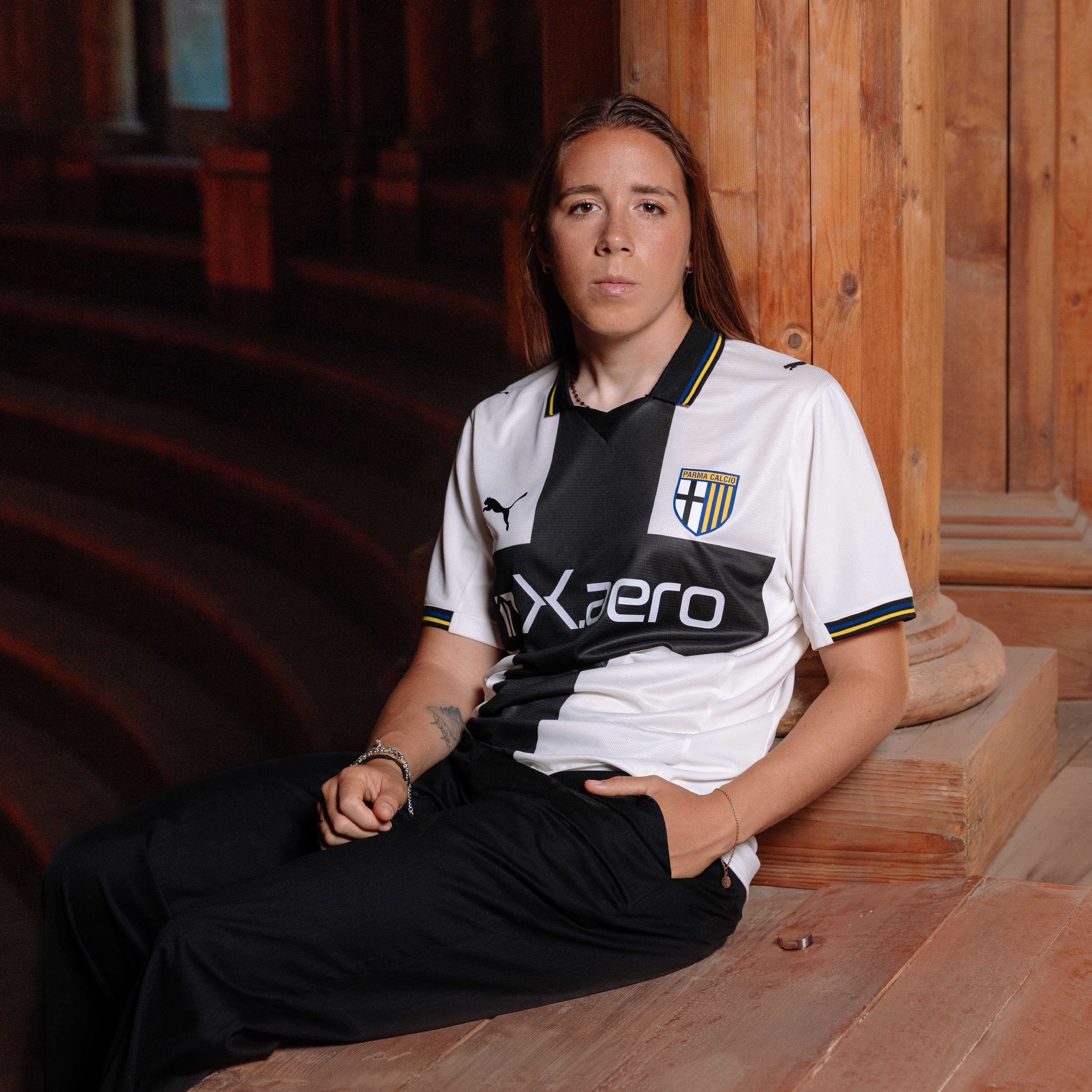 Calciatrice Ambrosi in posizione seduta con nuova maglia gara Home parma calcio bianca con croce nera e dettagli a righe gialloblu su colletto e maniche sospesa in aria con fondo teatro Farnese