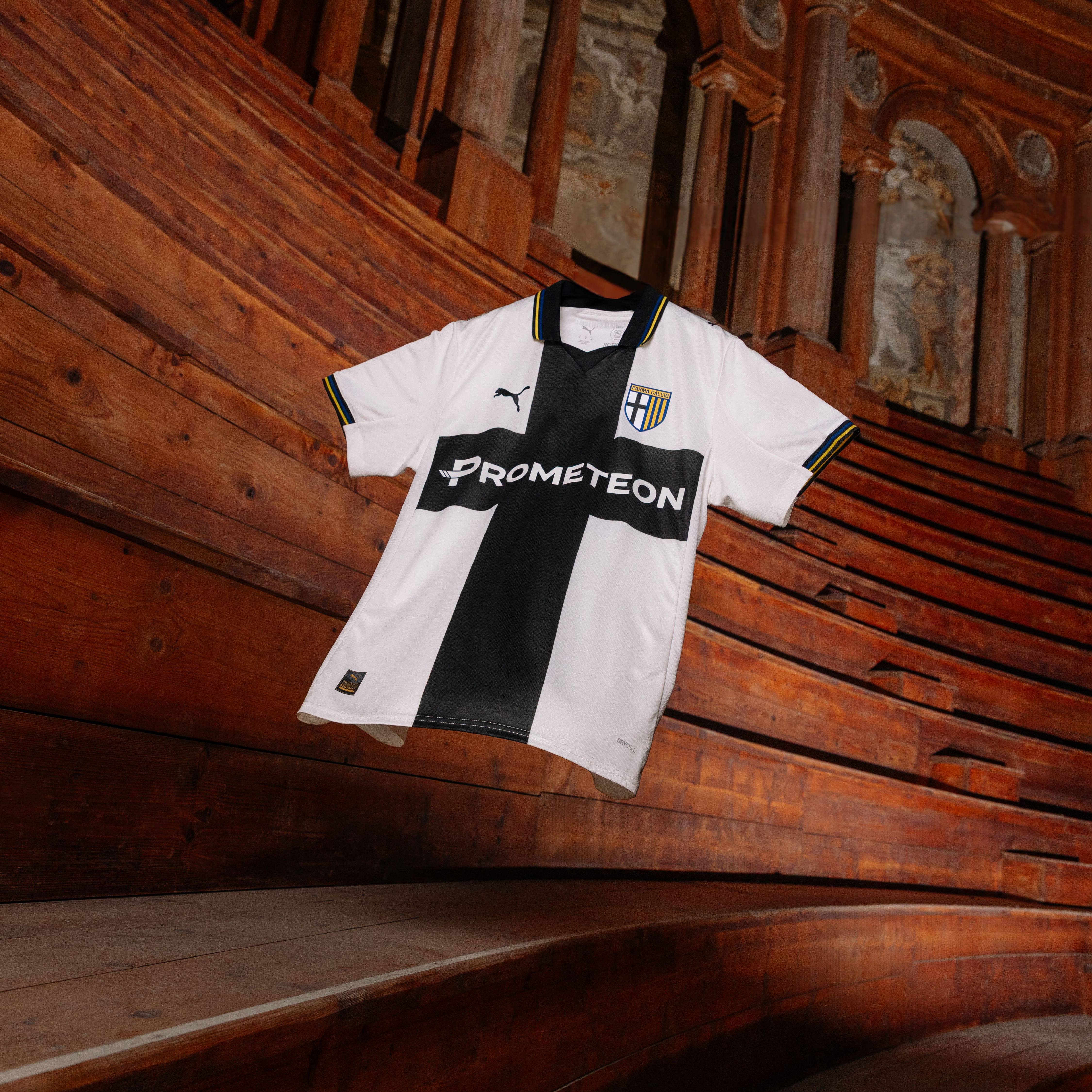 Maglia gara Home Parma Calcio maschile senza sponsor bianca con croce nera e dettagli a righe gialloblu su colletto e maniche sospesa in aria con fondo teatro Farnese